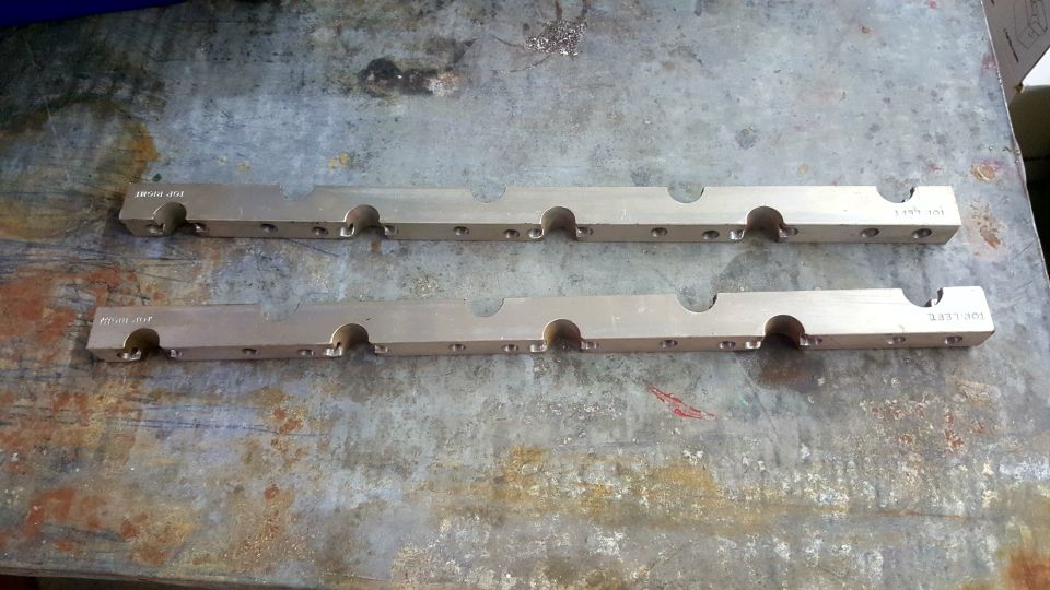 Two identical, long, flat, metallic engine parts with circular cutouts and small holes, lying on a textured metal surface