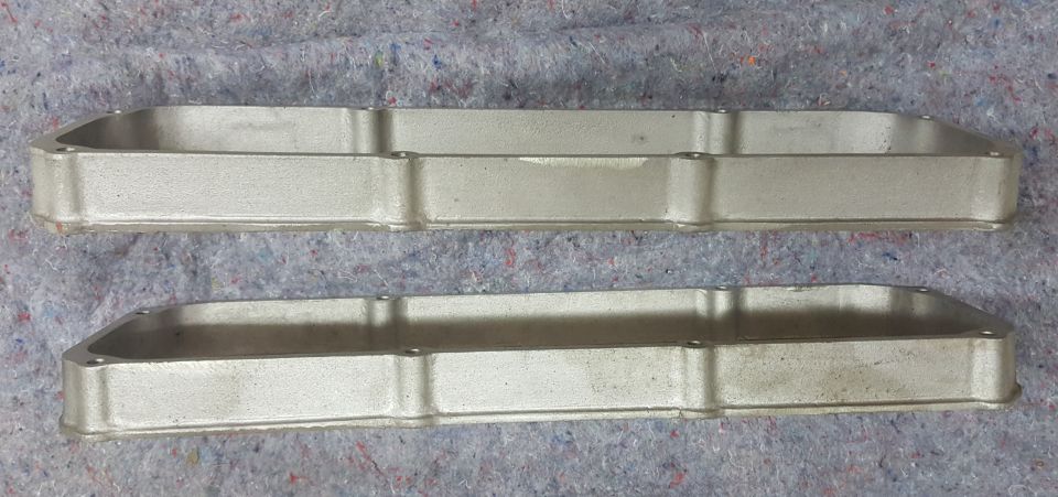 Pair of aluminum valve covers on a textured surface. Supermotors.net logo visible