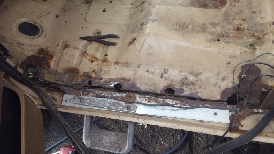Interior floor pan showing extensive rust and holes, with a pair of pliers on top