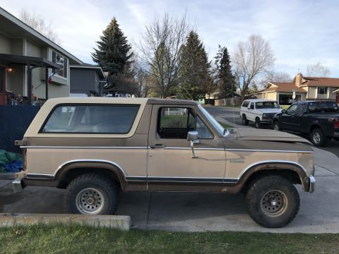 1985 Ford Bronco - A side view of a two-tone brown Ford Bronco parked in a driveway with other vehicles visible in the background.