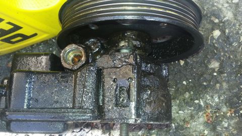 Greasy power steering pump with pulley attached. Yellow container with black text is in the background