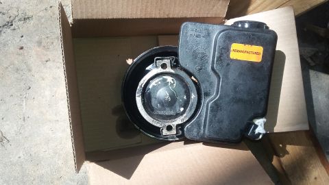 Black plastic power steering fluid reservoir with a yellow "REMANUFACTURED" sticker, inside a cardboard box