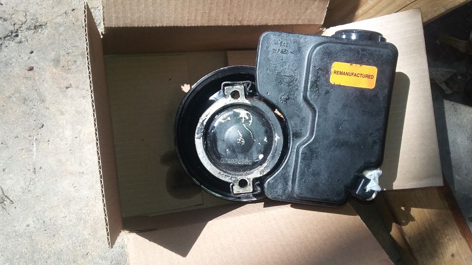 Black plastic power steering fluid reservoir with a yellow "REMANUFACTURED" sticker, inside a cardboard box