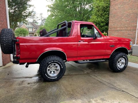 1994 Ford Bronco - The side profile of a red Ford Bronco is shown parked on a wet concrete driveway next to a brick building. The vehicle features off-road ti…