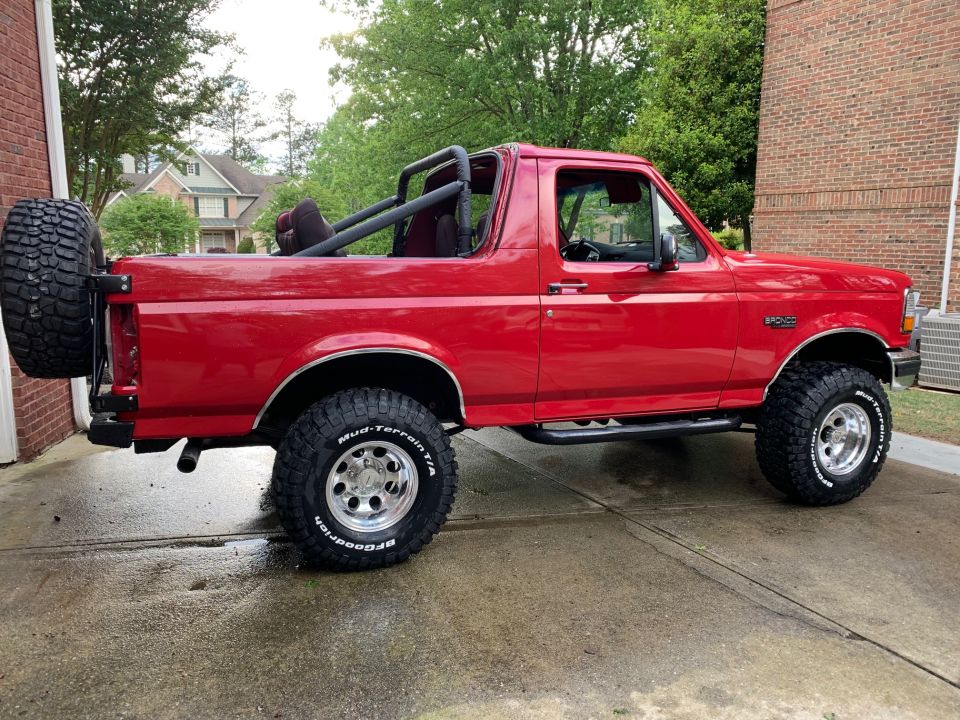 Red Ford Bronco parked on a wet driveway, side view, with off-road tires and roll cage.
