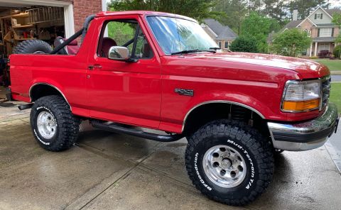 1994 Ford Bronco - A bright red, lifted Ford Bronco is parked on a wet concrete driveway in front of a residential garage. The vehicle features large BFGoodri…