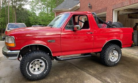 1994 Ford Bronco - Side view of a red Ford Bronco with aftermarket wheels and tires parked on a wet driveway in front of a brick house.