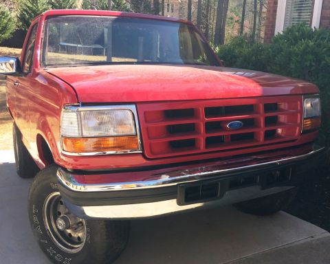 1994 Ford Bronco - The front view of a red Ford pickup truck is shown parked on a concrete driveway. The truck features a chrome bumper and grille with a blue…