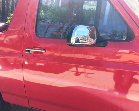 1994 Ford Bronco - The side of a red Ford Bronco is shown, featuring a chrome door handle and a chrome side mirror reflecting a street scene. The vehicle appe…