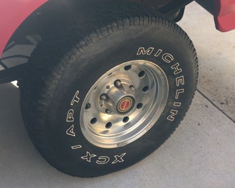 1994 Ford Bronco - A close-up shot shows the driver's side front tire and wheel of a red vehicle. The tire is a Michelin APT XCX, and the chrome wheel has a r…
