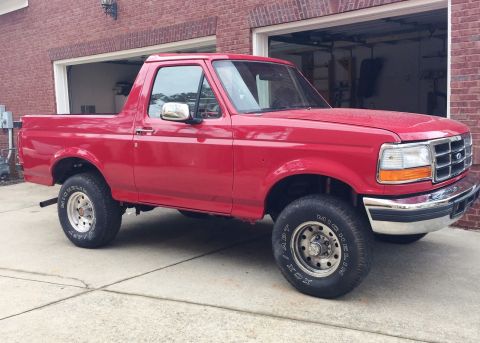 1994 Ford Bronco - A red 1990s Ford Bronco is parked in front of a brick garage. The SUV is shown from a 3/4 front angle, with its driver's side facing the ca…