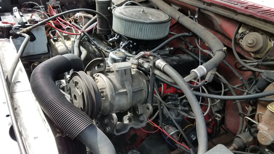 Engine bay of a red 1985 Ford Bronco with visible AC compressor, air filter, and wiring.
