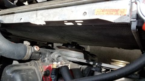 1985 Ford Bronco - A close-up view of the engine bay of a 1985 Ford Bronco, showing the radiator, pulleys, and a drive belt. A yellow sticker with red text is…