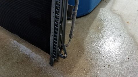1985 Ford Bronco - A close-up view of an air conditioning condenser unit, likely from a vehicle, resting on a wet concrete surface next to a blue container.