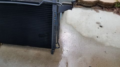 1985 Ford Bronco - A black automotive radiator or condenser unit is shown resting on a wet concrete surface. The unit has a dark gray metal bracket attached t…