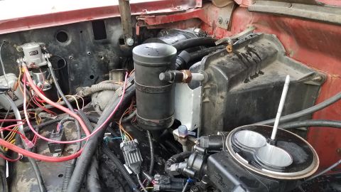 1985 Ford Bronco - The engine bay of a red 1985 Ford Bronco shows a complex wiring harness, a black cylindrical component with a brass fitting, and a throttle…