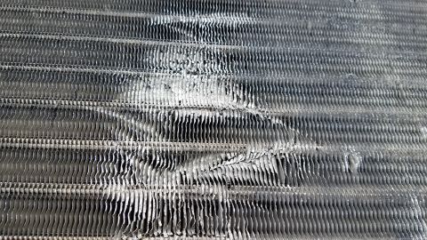 1985 Ford Bronco - A close-up, angled view of a damaged and dirty aluminum condenser coil, likely from an HVAC system. The fins are bent and clogged with debr…