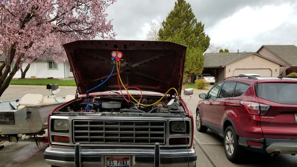 1985 Ford Bronco with hood open, connected to AC gauges, parked next to a red Ford Escape.