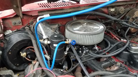 1985 Ford Bronco - The engine bay of a red Ford Bronco shows a modified air intake system and a black AC receiver/drier with blue refrigerant lines connected.