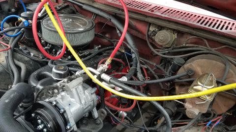 1985 Ford Bronco - A close-up view of the engine bay of a red Ford Bronco, focusing on the air conditioning system components and hoses. The image shows the A…