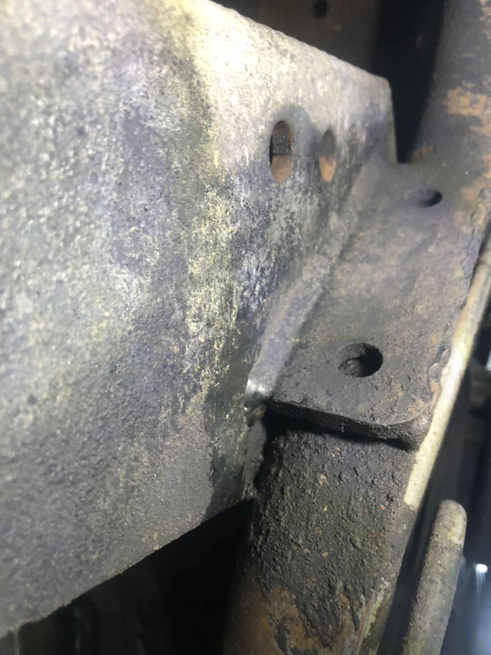 Close-up of a rusted, greasy metal bracket with bolt holes on a vehicle frame.