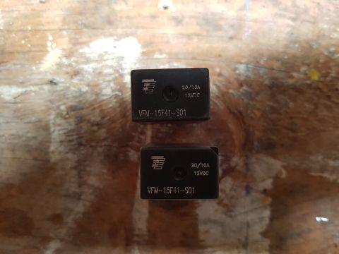 1978 Ford Bronco - Two black automotive relays are shown from a top-down perspective, resting on a wooden surface. Both relays have identical markings: a logo…