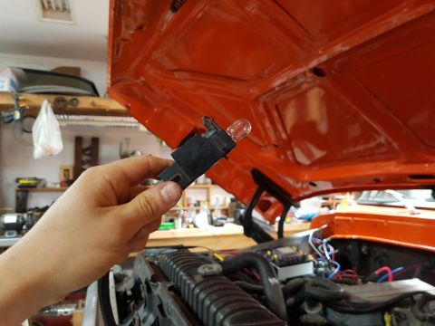 1978 Ford Bronco - A hand holds a light bulb socket and bulb, with the orange hood of a vehicle open in the background.