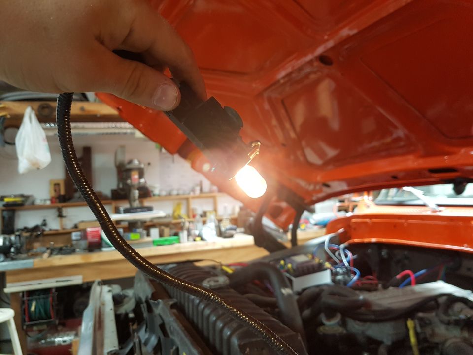 Hand holding a test light under the hood of an orange vehicle, showing engine components and wiring