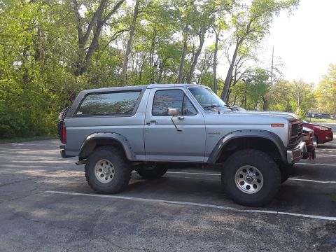 1985 Ford Bronco - This lifted Ford Bronco is parked in a lot with trees overhead.