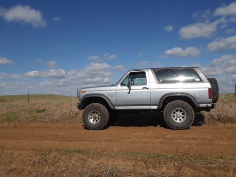 1985 Ford Bronco - A silver Ford Bronco from the early 1980s sits on a dirt road under a partly cloudy sky.