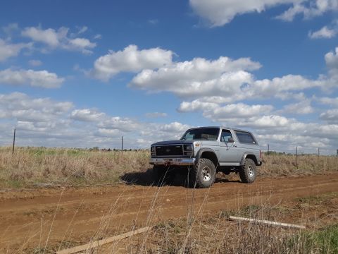 1985 Ford Bronco - A silver 1985 Ford Bronco sits on a dirt road, its stance accentuated by the open field and dramatic clouds.