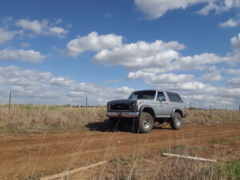 Silver 1985 Ford Bronco parked on a dirt road under a cloudy sky