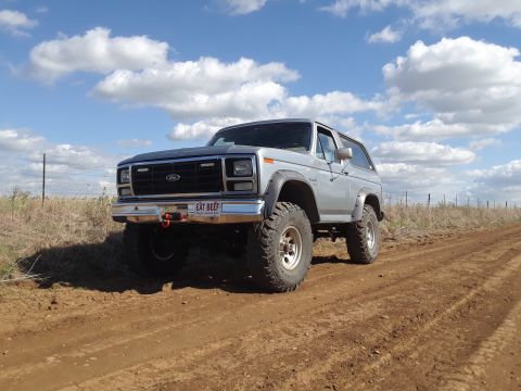 1985 Ford Bronco - Silver 1985 Ford Bronco with off-road tires driving on a dirt path under a cloudy sky.