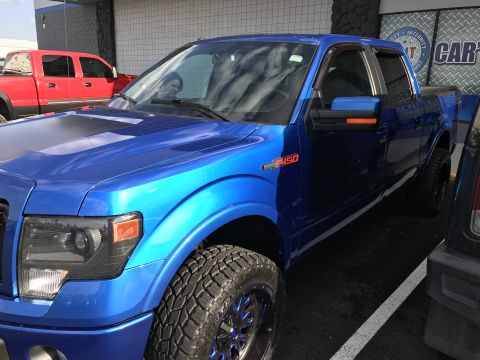2013 Ford F-150 - A blue 2013 Ford F-150 is parked in a lot, with a red truck visible in the background.