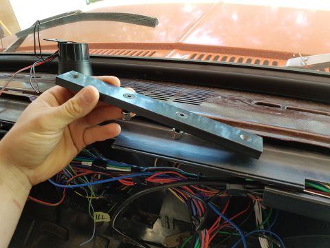 1978 Ford Bronco - A hand holds a dark rectangular bracket with two visible screw holes over a car's dashboard, revealing a complex wiring harness underneath.