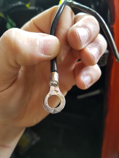 1978 Ford Bronco - A hand holds a black wire with a ring terminal attached. The ring terminal is silver and shows signs of wear and tear.