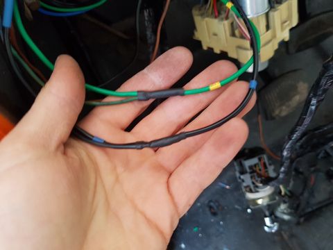 1978 Ford Bronco - A hand holds two black wires with heat shrink tubing and blue and yellow tape markings. In the background, a complex wiring harness and fus…