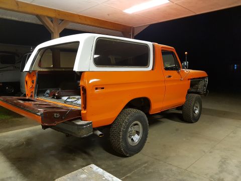 1978 Ford Bronco - An orange 1978 Ford Bronco with a white roof is parked inside a garage with the tailgate down. The vehicle is missing its front fenders and…