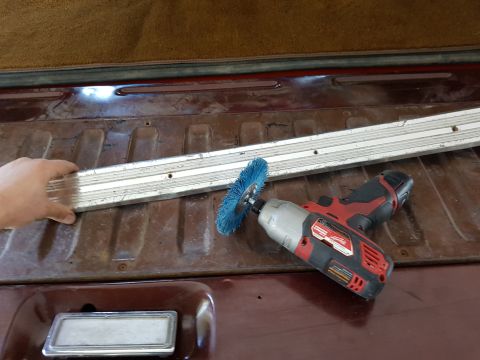 1978 Ford Bronco - A person's hand holds a metal trim piece above the bed of a dark red pickup truck. A red and black power tool with a blue brush attachment…