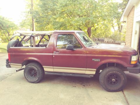 1996 Ford Bronco - The side view of a maroon Ford Bronco with a tan lower body trim and a soft top is parked on a concrete driveway. A spare tire is mounted v…
