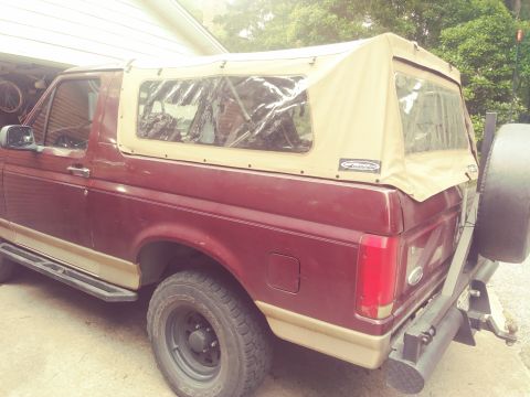 1996 Ford Bronco - The rear passenger side of a maroon Ford Bronco with a tan soft top is shown parked outdoors. A spare tire is mounted on the rear bumper, a…