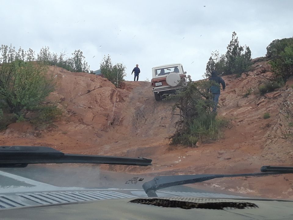 Classic Ford Bronco climbing a steep, rocky trail with two people nearby.
