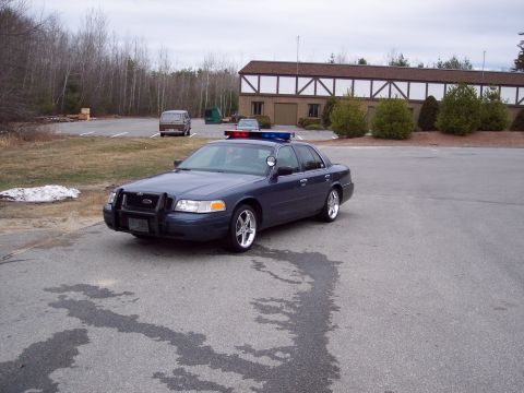 2001 Ford Crown Victoria - Three-quarter front view of a dark blue-gray Ford Crown Victoria police-package sedan parked on an empty asphalt lot, fitted with a black t…