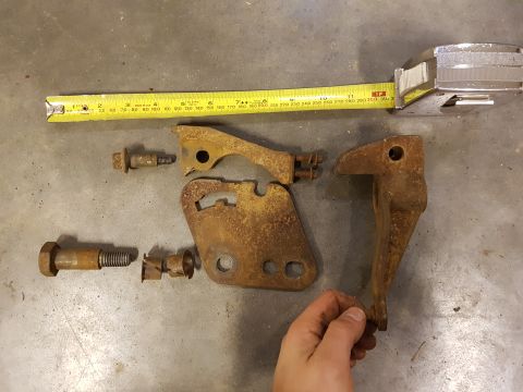 1978 Ford Bronco - Several rusty metal brackets and bolts are laid out on a concrete floor next to a yellow measuring tape and a chrome tape measure. A hand i…