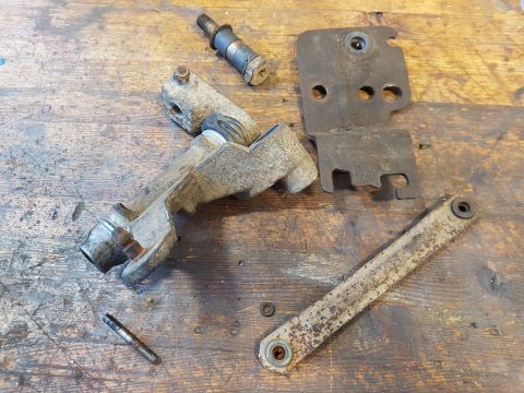 1978 Ford Bronco - A collection of disassembled metal automotive parts, including a cast metal bracket with a pivot mechanism, a rusted metal plate, and a lon…