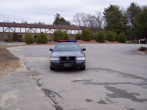 2001 Ford Crown Victoria - Police car parked in front of a building with trees and a cloudy sky.
