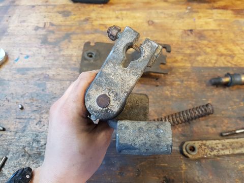 1978 Ford Bronco - A hand holds a rusty metal automotive part, possibly a linkage or bracket, against a wooden workbench. Other metal parts and a spring are v…