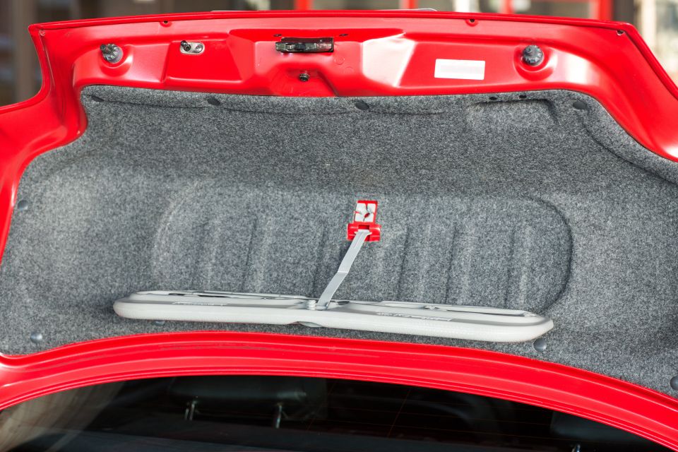 Open red car trunk interior with grey carpet and a white object secured by a red clip.