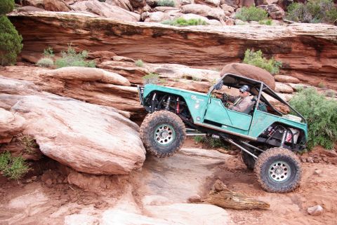1995 Ford Bronco - A modified teal Ford Bronco is navigating a steep, rocky incline in a desert environment. The vehicle is angled, with its front passenger-s…