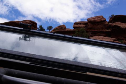 1995 Ford Bronco - A close-up view of a vehicle's windshield showing a cracked surface with reflections of red rock formations and a blue sky with clouds. A b…